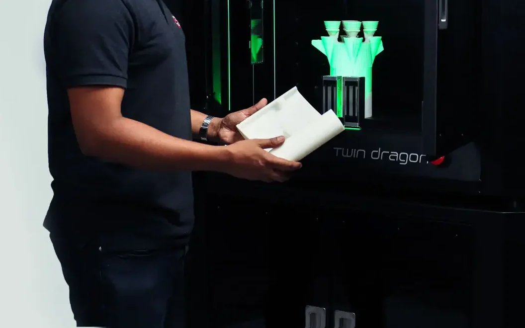 A Fracktal Works engineer inspecting a finished 3D printed part beside a Twin Dragon printer running a build